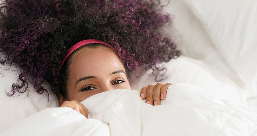 Mujer tumbada en la cama tapándose la mitad de su cara y sonriendo